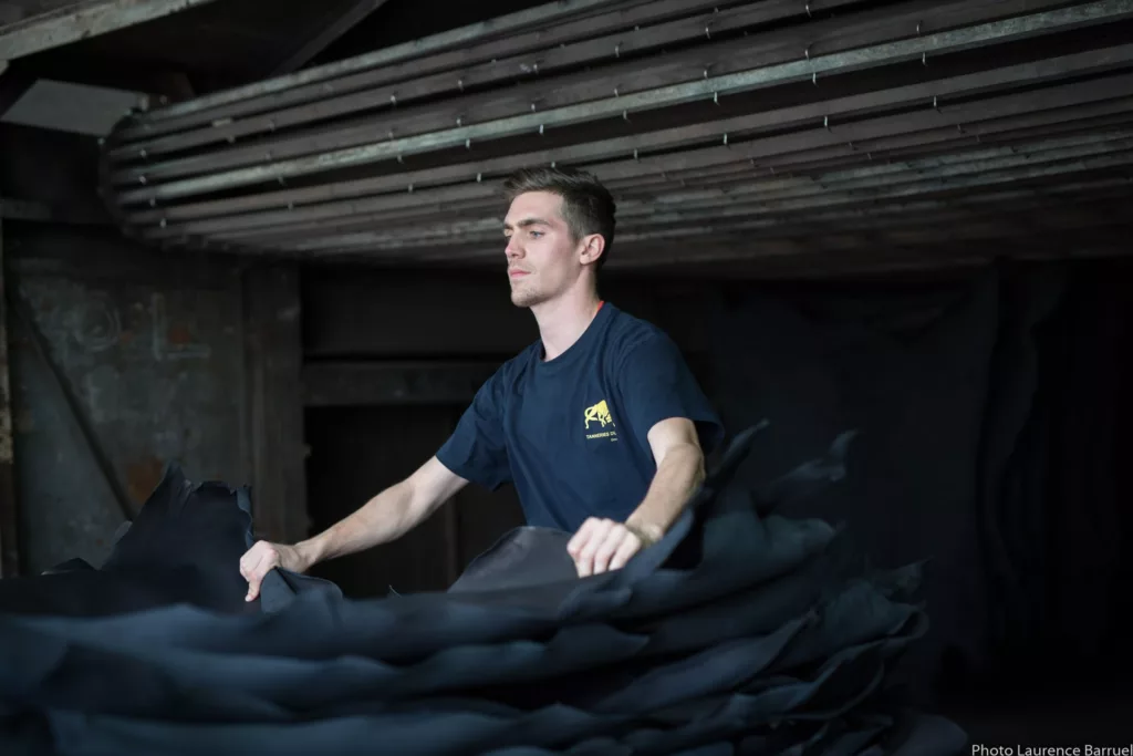 A man in a blue T-shirt handles a pile of black leather in the dark atmosphere of a tannery