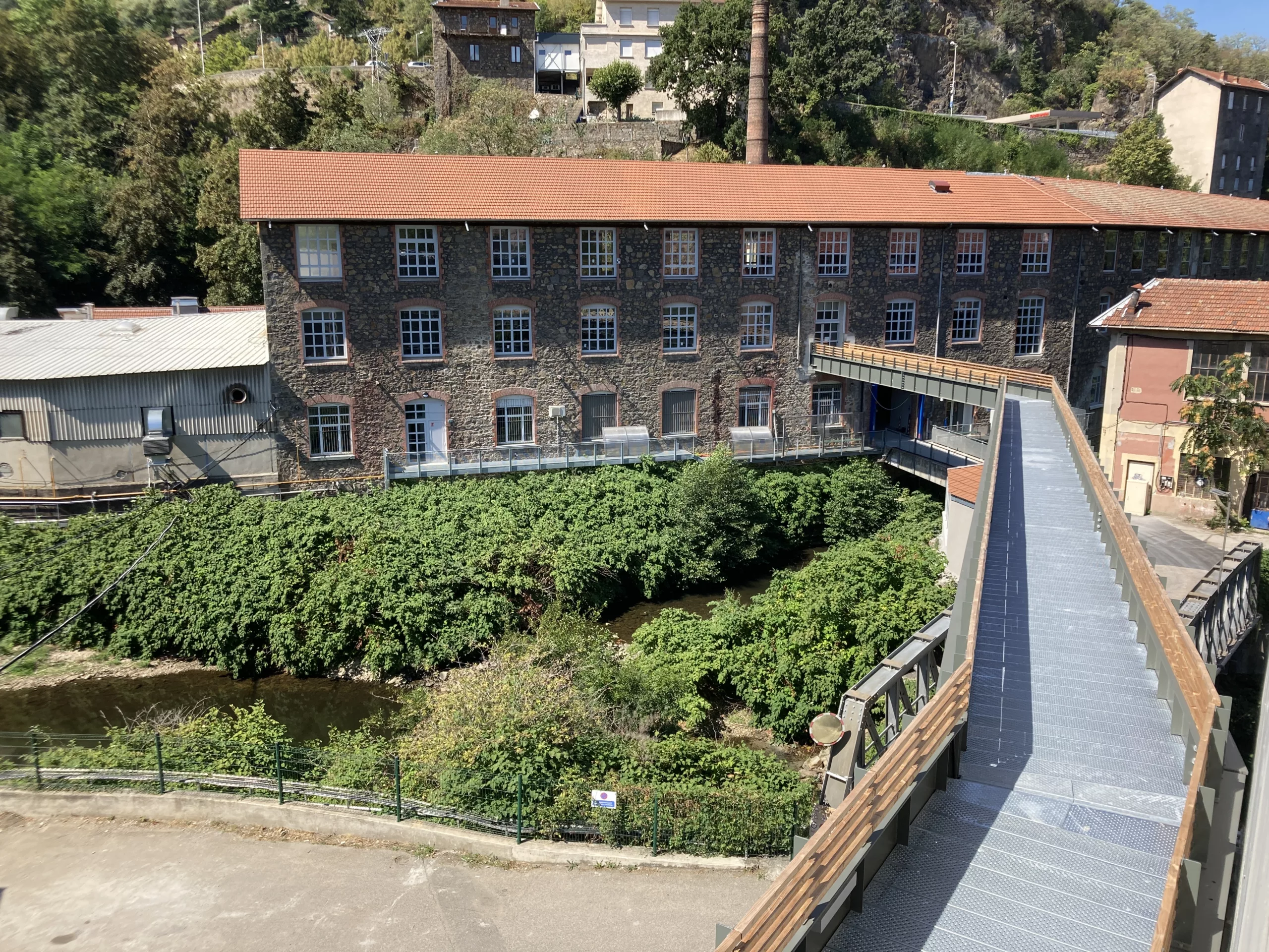 Vue extérieure d’un bâtiment en briques entouré de verdure, avec passerelle métallique menant à l’entrée.