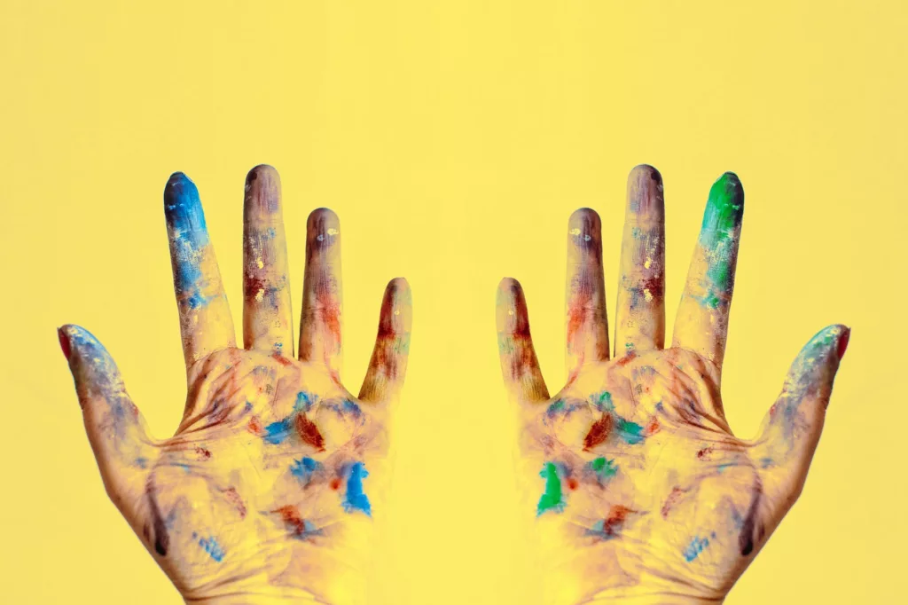 Two hands, webbed upwards, extensively covered in brightly colored paint on a yellow background.