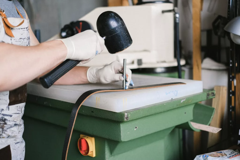 A craftsman uses a mallet to punch a strip of leather on a work surface. White gloves protect his hands. The craft workshop is visible in the background.