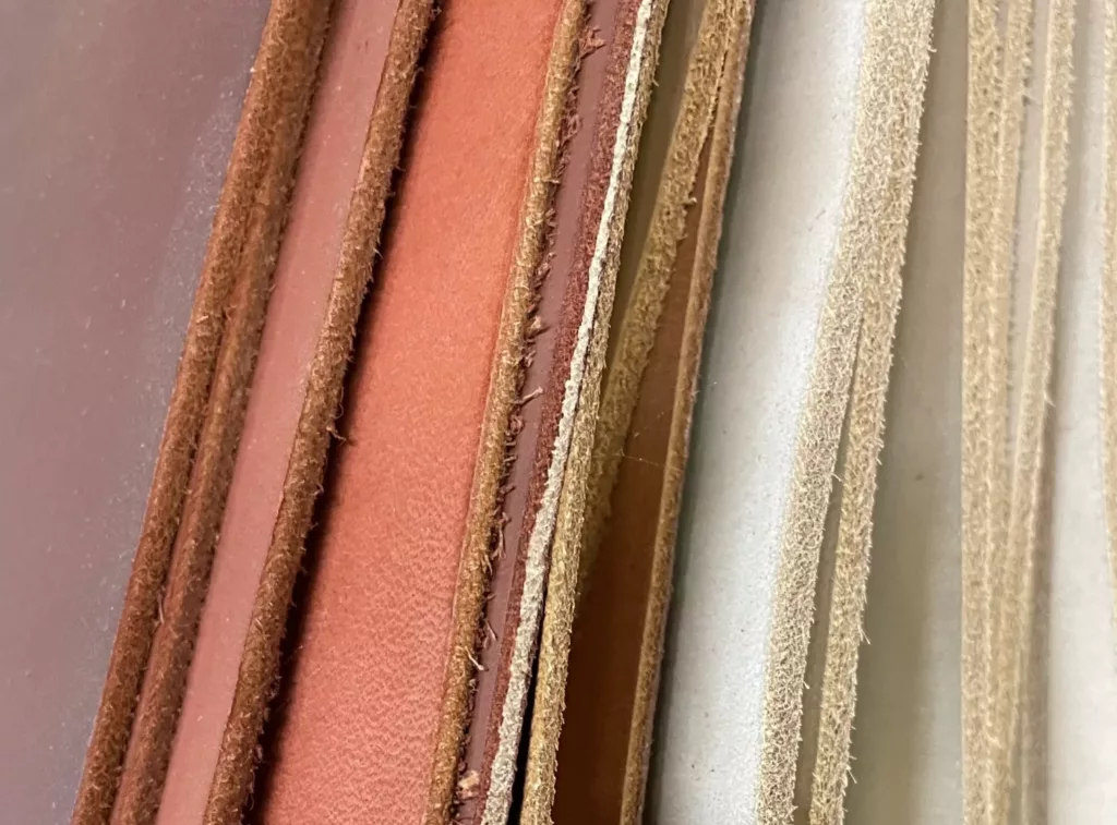 The image displays several cowhide leather strips arranged side by side. The strips vary in color and shade, ranging from earthy reddish and dark brown tones to light beige and almost white hues. They appear to be of similar width and have a smooth, uniform texture. The edges are clean, suggesting they have been neatly cut. The color variations likely indicate different stages of the tanning process or leather from different animals. The arrangement suggests a leather display, possibly in a leather goods store or a leatherworking workshop.