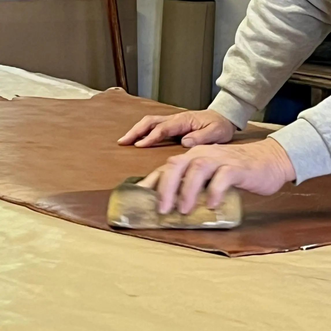 Person using a wooden sponge to smooth and polish a brown cowhide, with visible sponge marks on the surface.