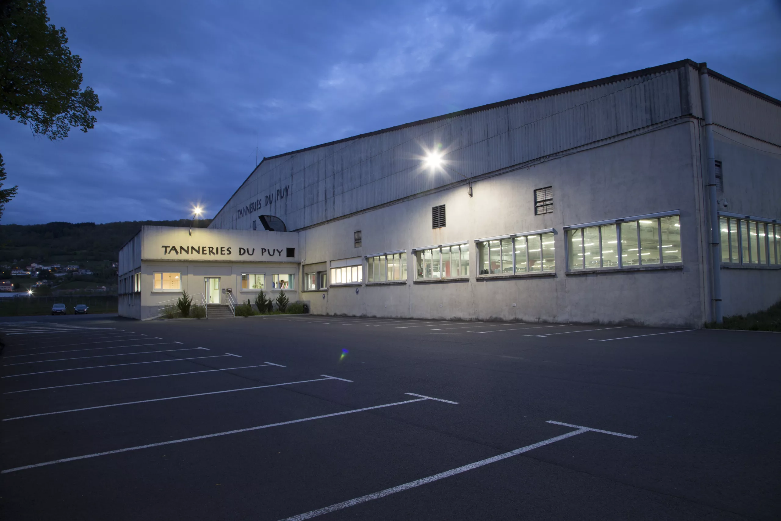 Une photo des Tanneries du Puy, montrant un grand bâtiment industriel avec des fenêtres éclairées et un parking vide au crépuscule.